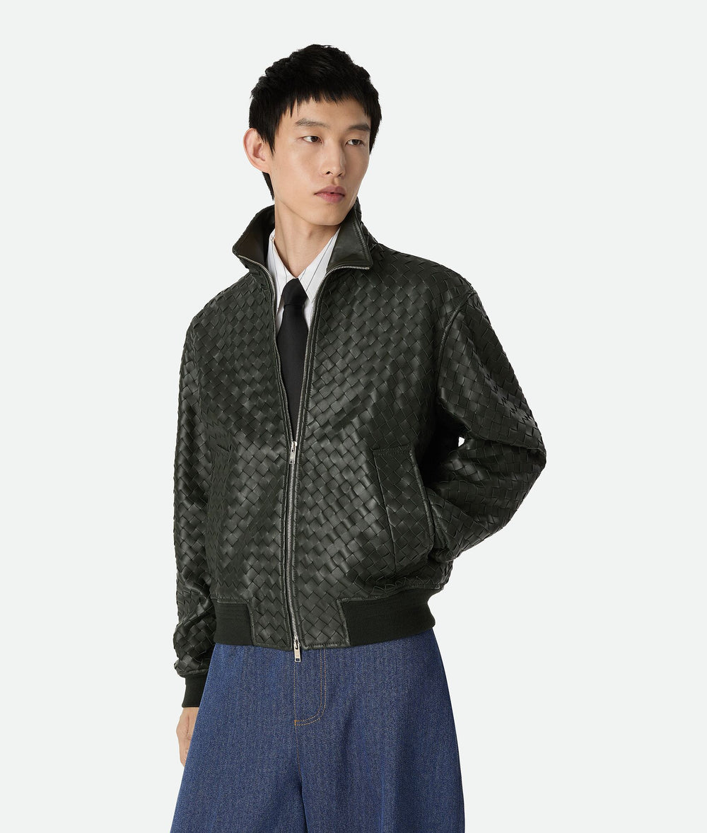 Dark green jacket worn open: interlaced panels across front and sleeves, zipper, ribbed cuffs and hem; no view of hidden backing.