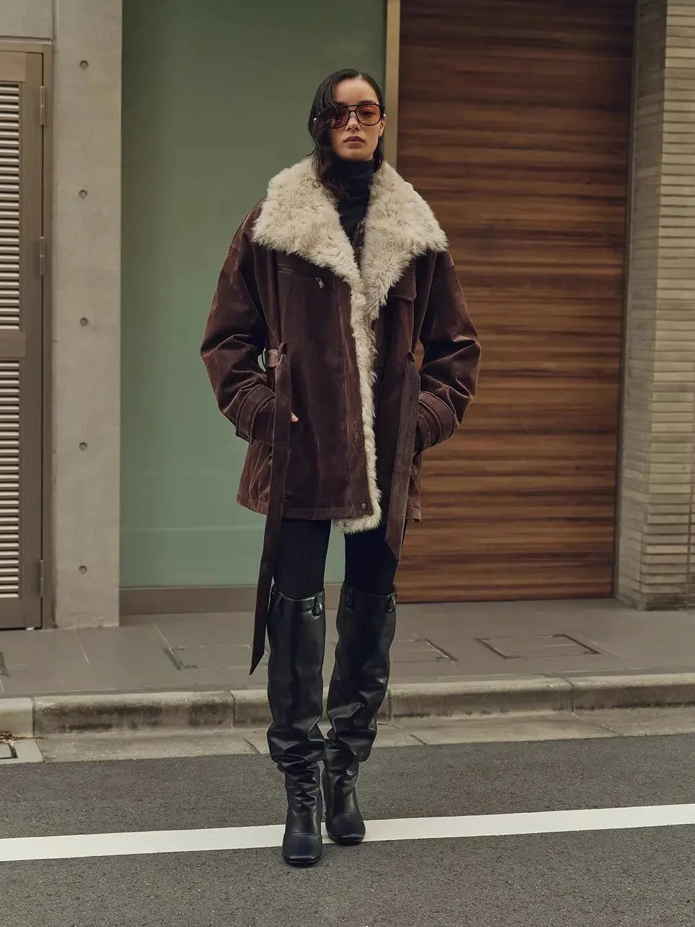 Brown Glacier Traveler parka with a pale collar over narrow dark trousers tucked into tall black boots