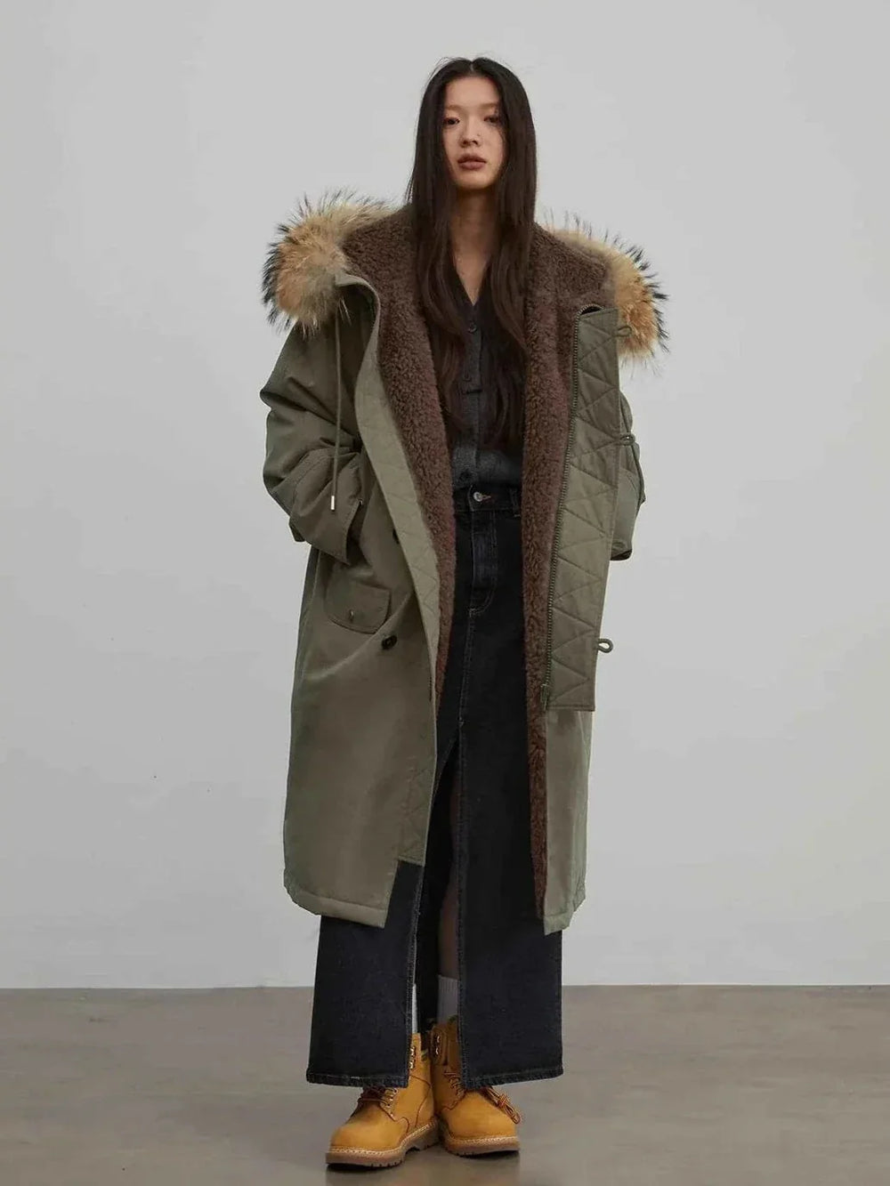 Olive Manchester parka worn open over a dark base and wide dark trousers with tan ankle boots