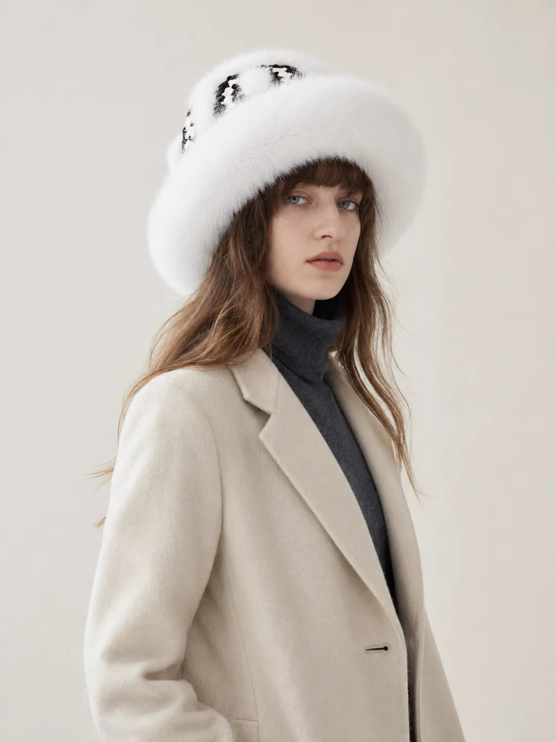 A woman wearing a wide-brimmed white fur hat with black sequins, a beige wool coat, and a dark gray turtleneck sweater, photographed against a plain light beige studio background.