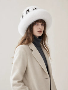 A woman wearing a wide-brimmed white fur hat with black sequins, a beige wool coat, and a dark gray turtleneck sweater, photographed against a plain light beige studio background.