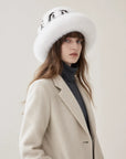 A woman wearing a wide-brimmed white fur hat with black sequins, a beige wool coat, and a dark gray turtleneck sweater, photographed against a plain light beige studio background.