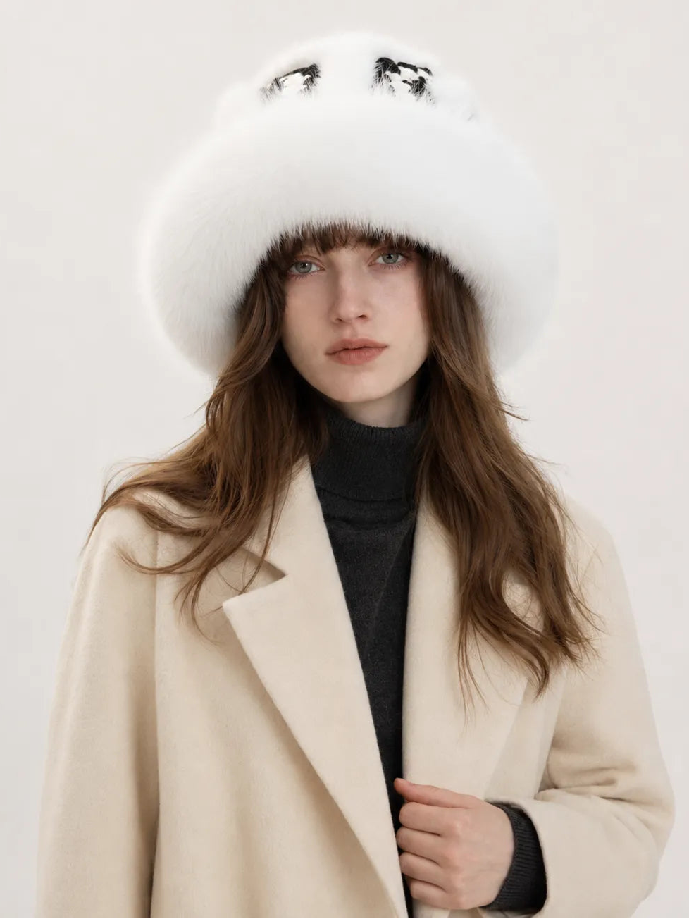 A woman wearing a large white fur hat with black embroidered eyes and a cream-colored coat over a black turtleneck, photographed against a plain light background.
