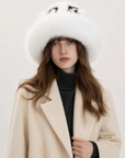 A woman wearing a large white fur hat with black embroidered eyes and a cream-colored coat over a black turtleneck, photographed against a plain light background.